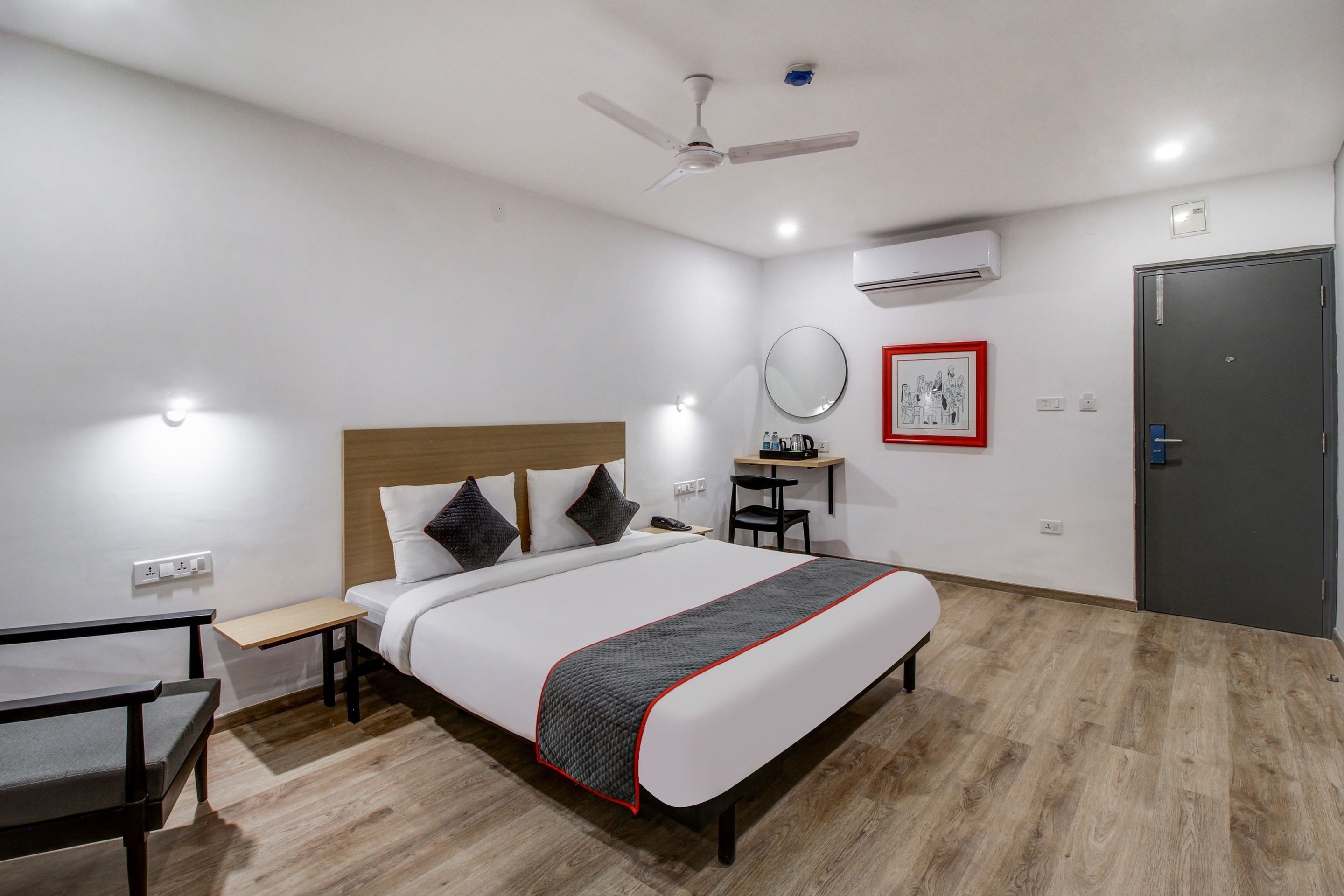 Een hotelkamer met een bed, nachtkastje, stoel, bureau, spiegel, kunstwerk en een airconditioning.