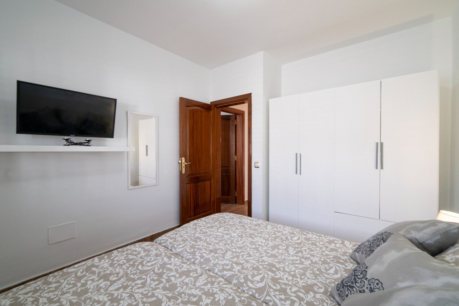 A bedroom with a bed, a wardrobe, a television mounted on the wall, and a door leading to another room.