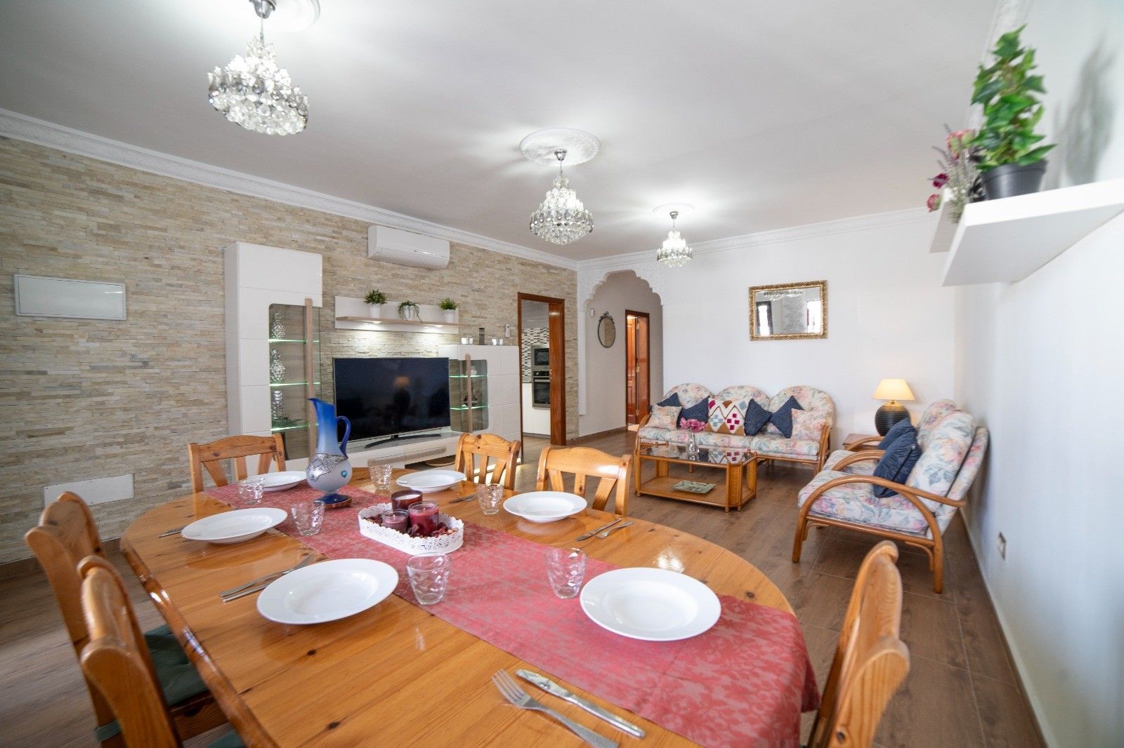 Un comedor con una mesa de madera dispuesta para cuatro personas, rodeada de sillas. Hay un televisor sobre un mueble bajo, un sofá con cojines y una planta en maceta sobre una estantería.