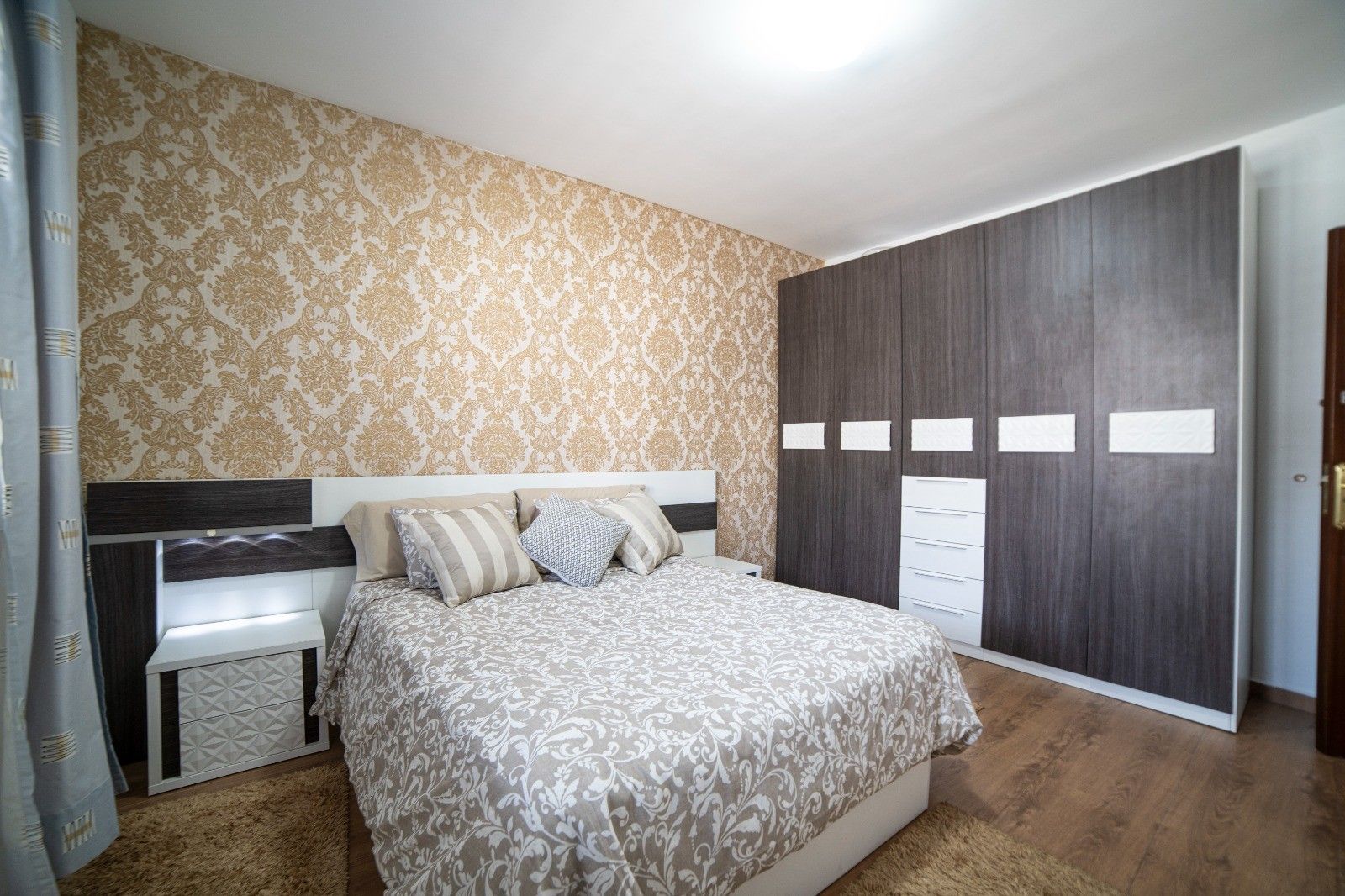 Un dormitorio con cama, mesitas de noche, armario y papel tapiz estampado.