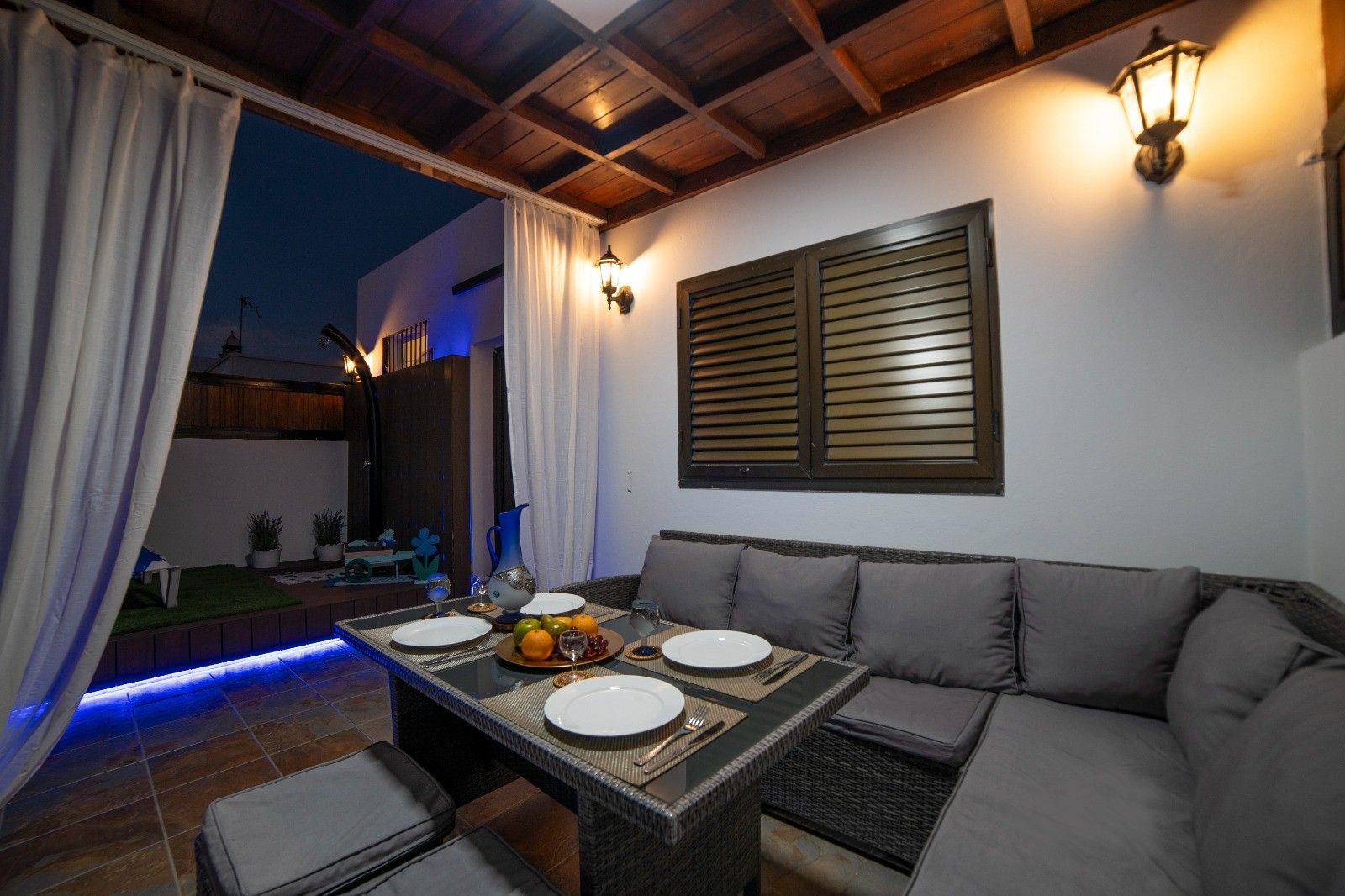 An outdoor dining area with a table set for two, surrounded by cushioned seating, a wall-mounted light fixture, and a window with shutters.