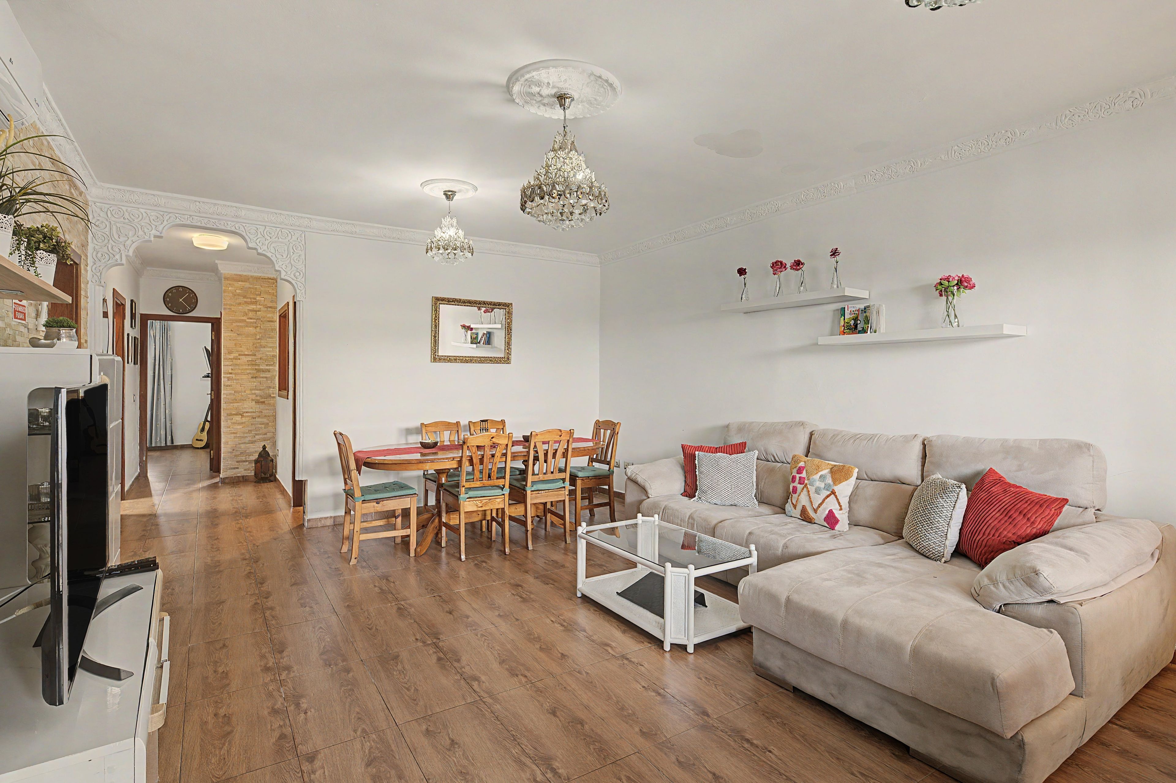 Un salon comprenant une table à manger en bois, des chaises, une table basse en verre, un canapé beige agrémenté de coussins et des étagères décoratives ornées de fleurs.