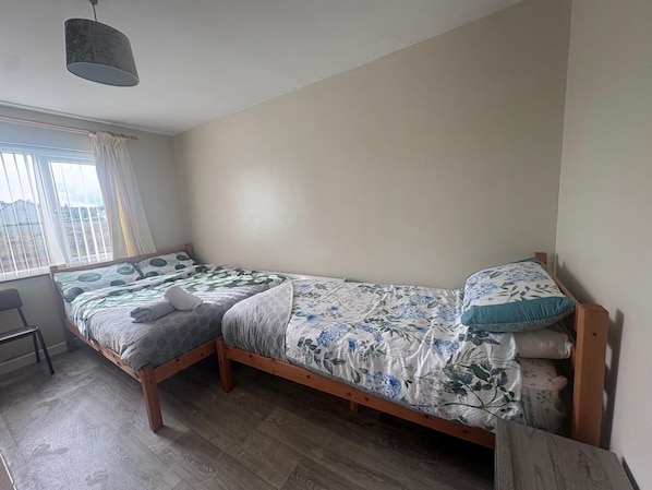 Una cama individual con colcha estampada y dos almohadas, piso de madera, ventana con cortinas y una lámpara colgante.