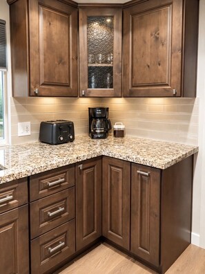 A kitchen with wooden cabinets, a granite countertop, a toaster, a coffee maker, and a window with blinds.