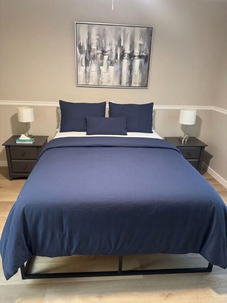 Un lit recouvert d’une couvre-lit bleu foncé et agrémenté d’oreillers assortis, flanqué de deux tables de chevet sur lesquelles reposent des lampes, dans une pièce aux murs de couleur neutre et où un tableau abstrait encadré est accroché au-dessus du lit.