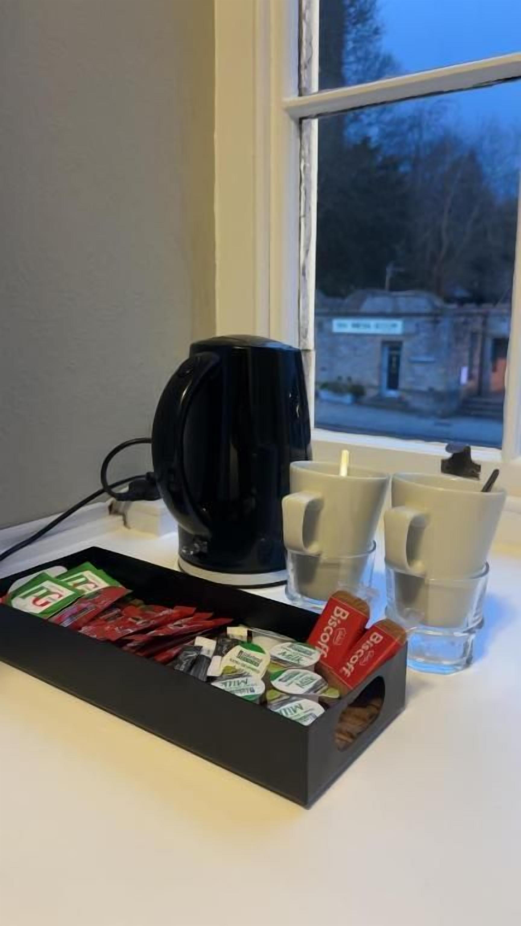 Una cafetera negra, dos tazas y una caja de bolsitas de té sobre una mesa junto a una ventana.
