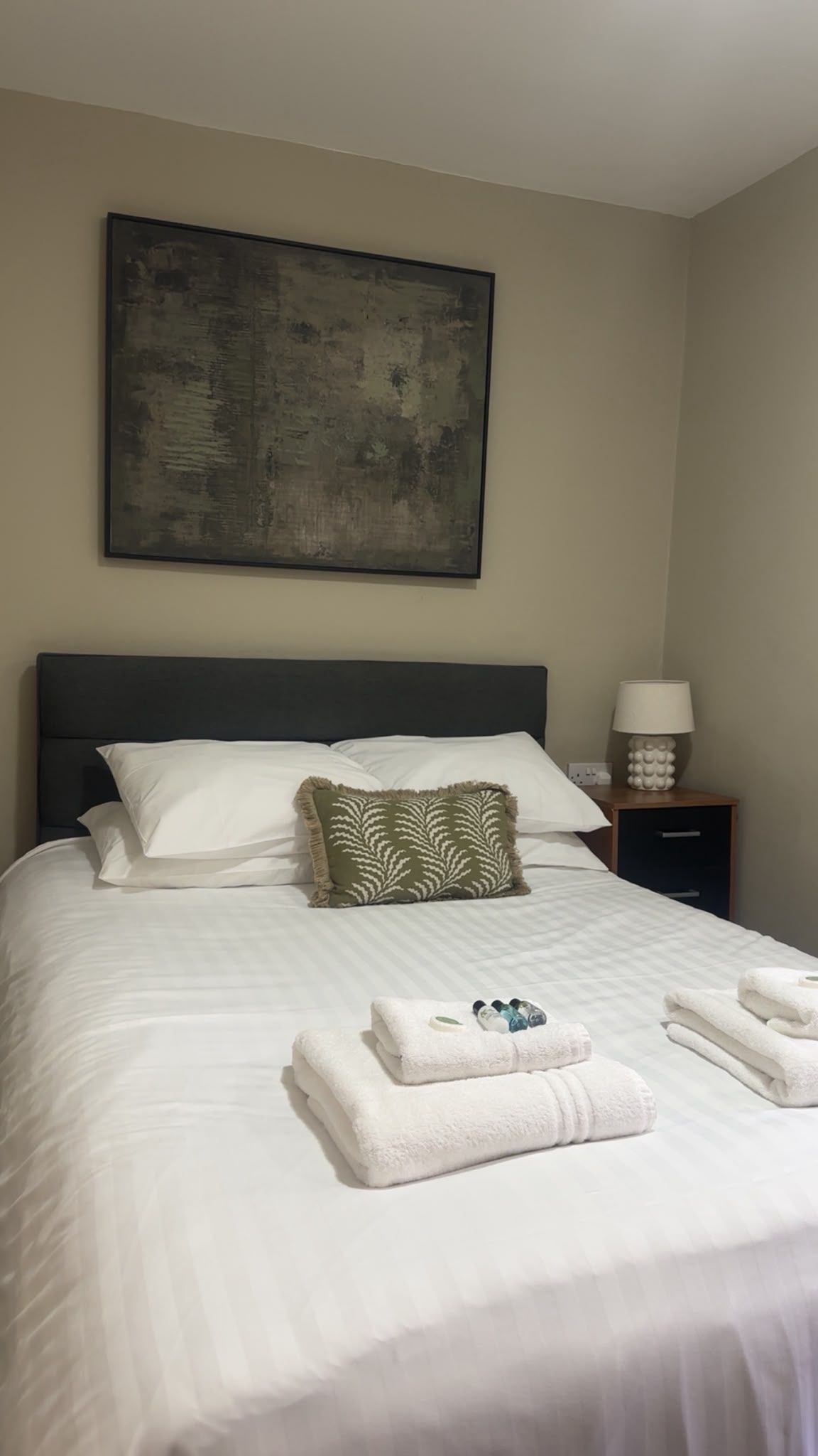 Una cama bien hecha con sábanas blancas y un cojín estampado en verde, una lámpara de noche y un cuadro en la pared.