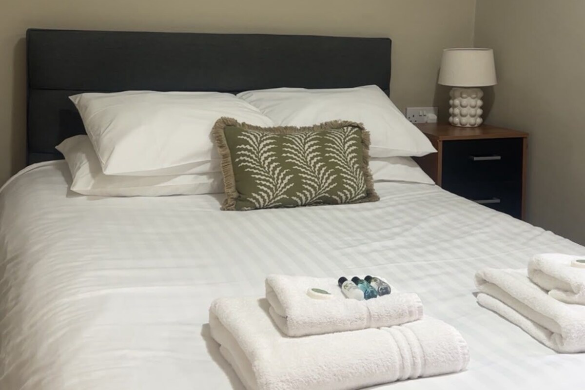 A neatly made bed with white linens and a green patterned pillow, a bedside lamp, and a painting on the wall.