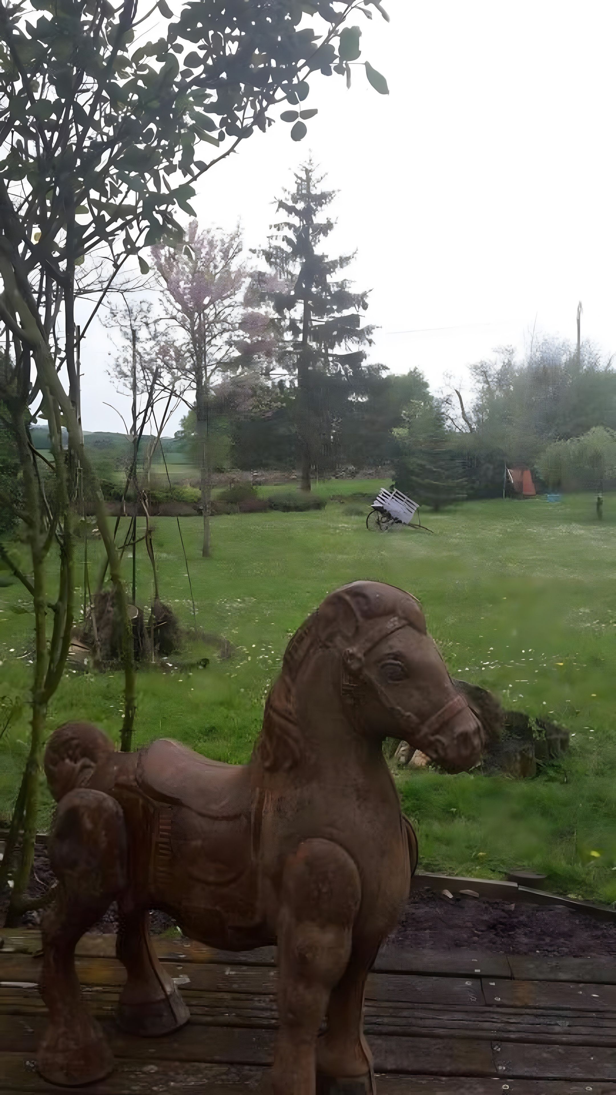 Eine Holzpferdskulptur in einer grünen Parklandschaft mit Bäumen und einem entfernten Gebäude.