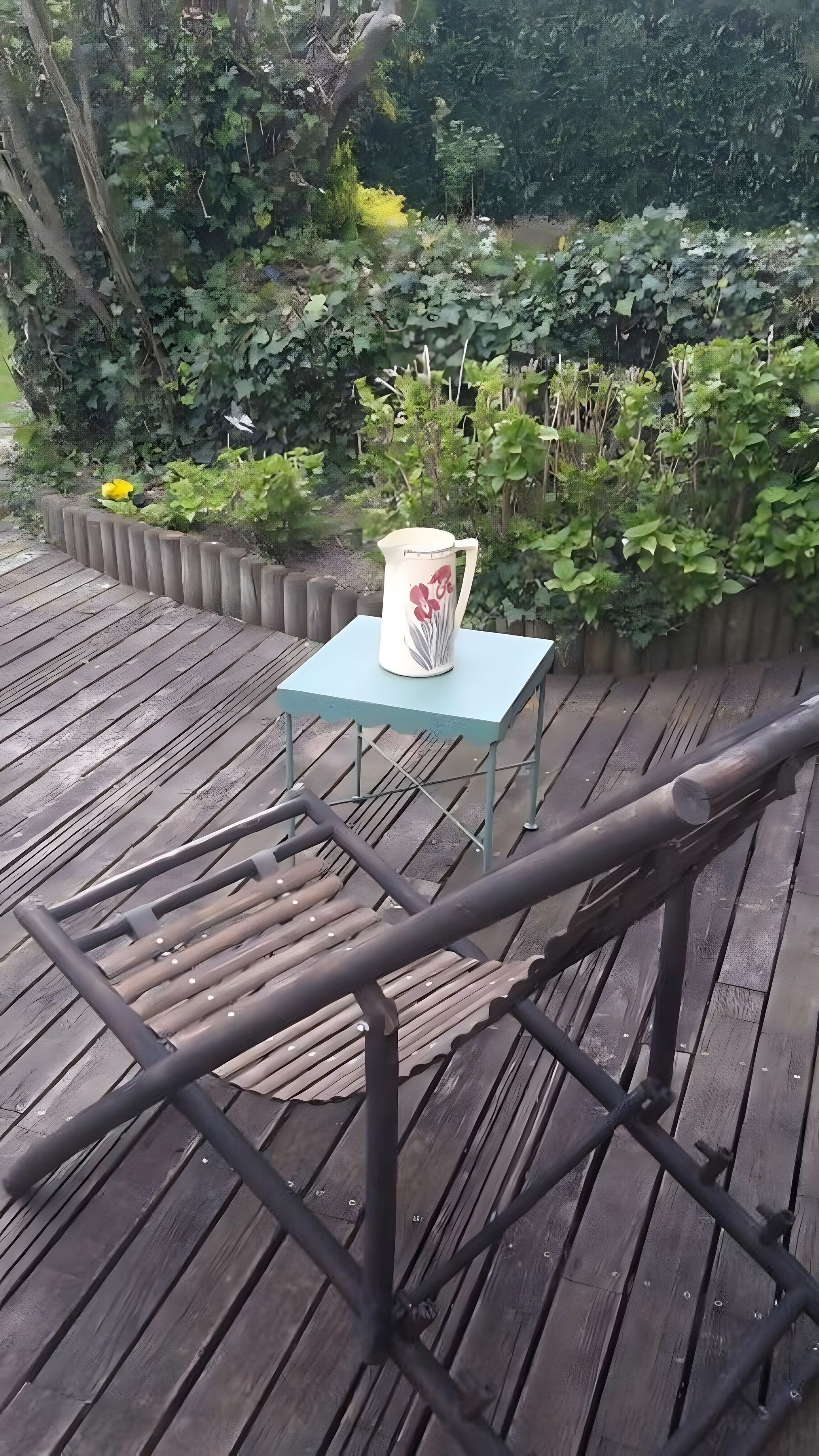 Un terrazza in legno con ringhiera metallica, un tavolino con un vaso decorativo e vegetazione lussureggiante sullo sfondo.