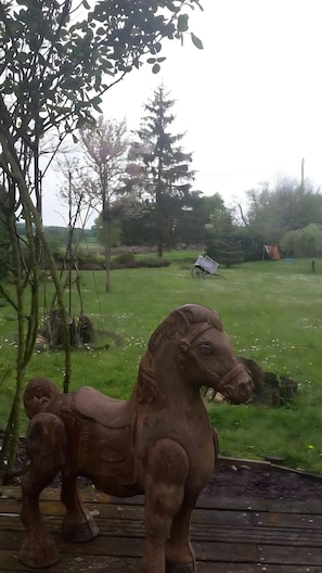 Eine Holzpferdskulptur in einer grünen Parklandschaft mit Bäumen und einem entfernten Gebäude.