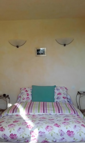 Ein Bett mit blumigem Bettzeug, einem türkisblauen Kissen und einem gerahmten Bild an der Wand.