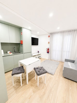 Una sala moderna con un sofá gris, una mesa de comedor blanca y dos taburetes grises acolchados, un televisor de pantalla plana montado en la pared y una alfombra gris sobre el suelo de madera.