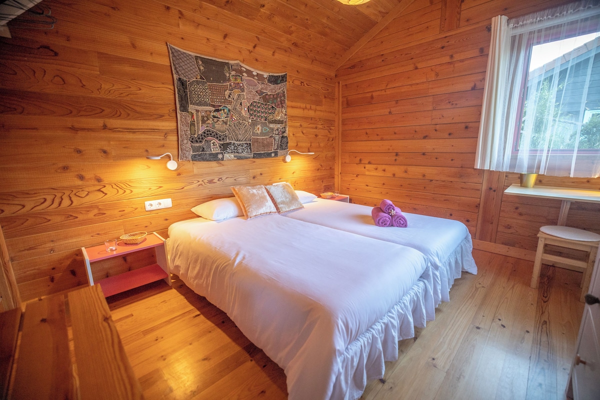 A wooden cabin room with two beds, a wall tapestry, and a window with curtains.