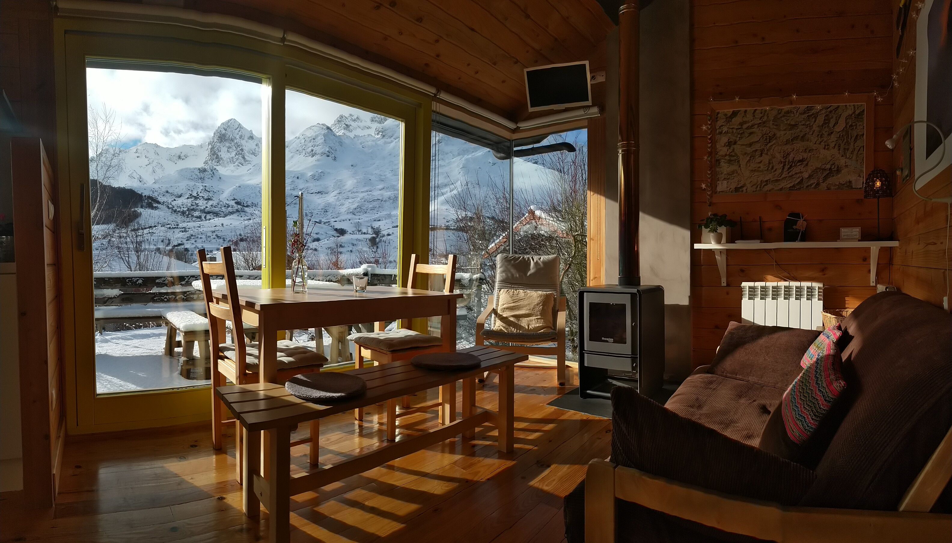 Una habitació acollidora amb interior de fusta, una taula, cadires, una panxa, un sofà, una llar de foc i vista a muntanyes cobertes de neu.