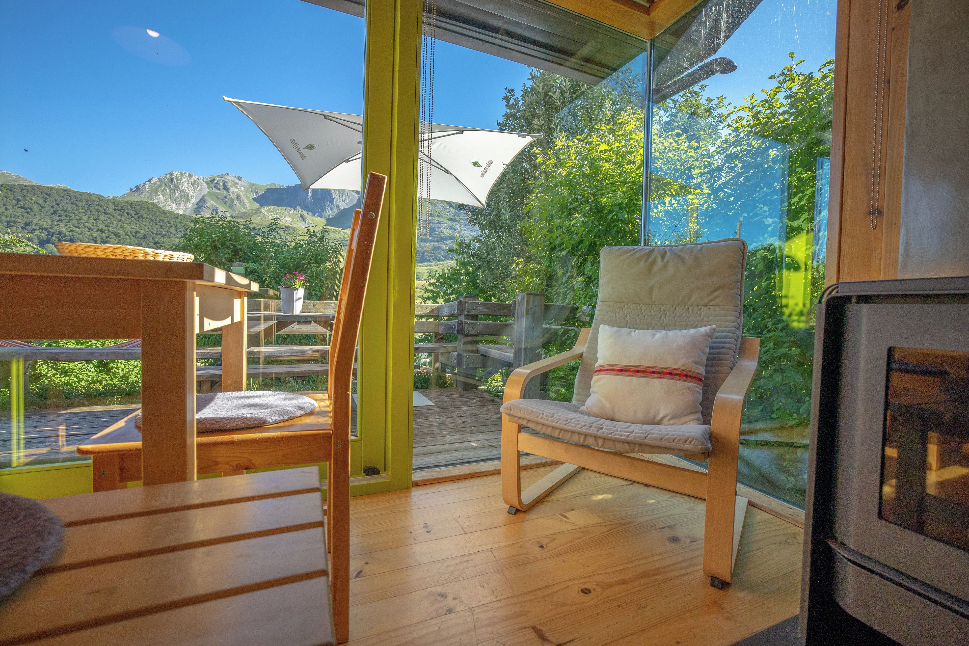 A wooden deck with a chair and table, a glass door leading to a mountain view, and a wooden structure with a white umbrella.
