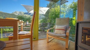 Une terrasse en bois avec une chaise et une table, une porte vitrée donnant sur une vue sur les montagnes, et une structure en bois surmontée d’un parasol blanc.