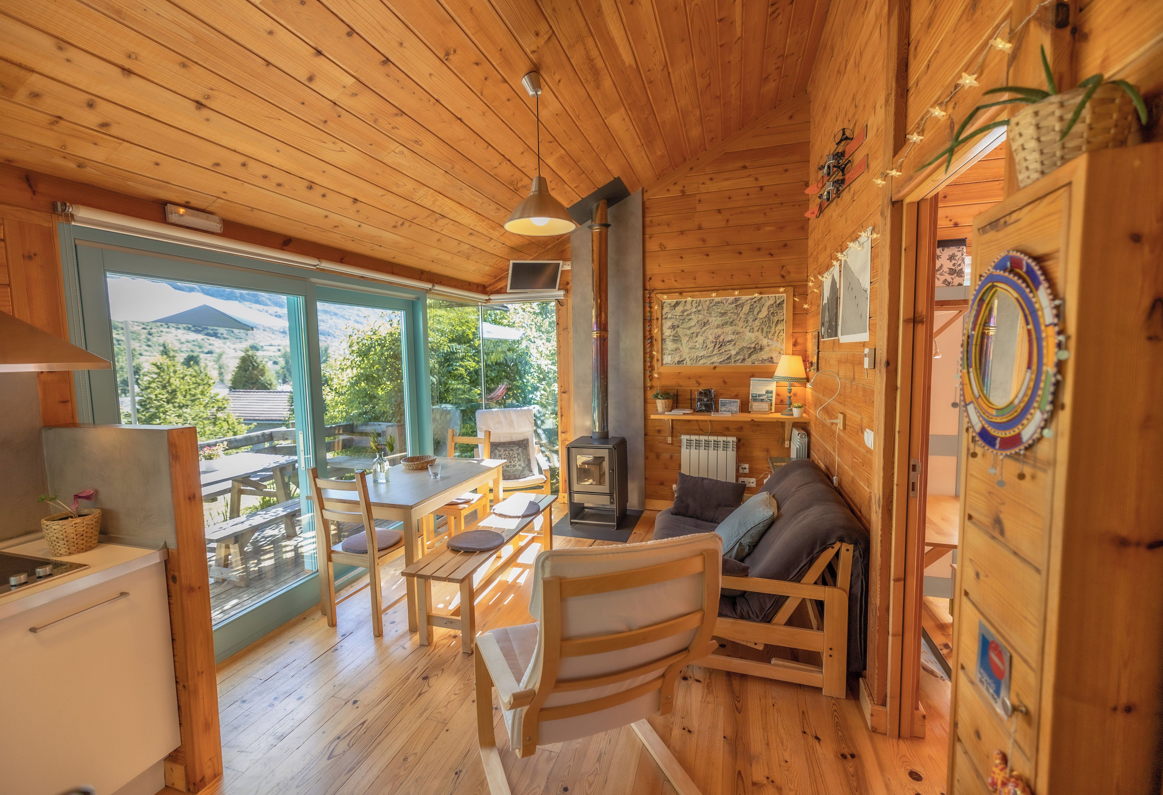Interior de una cabaña de madera con comedor, asientos, chimenea y vistas amplias a través de ventanas grandes.