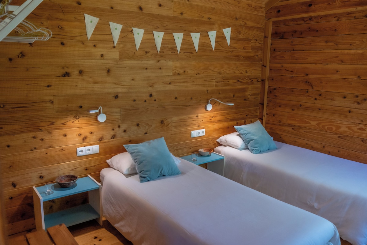 A wooden room with two beds, each with a pillow and a nightstand. There are wall-mounted lights and a power outlet on the wall.