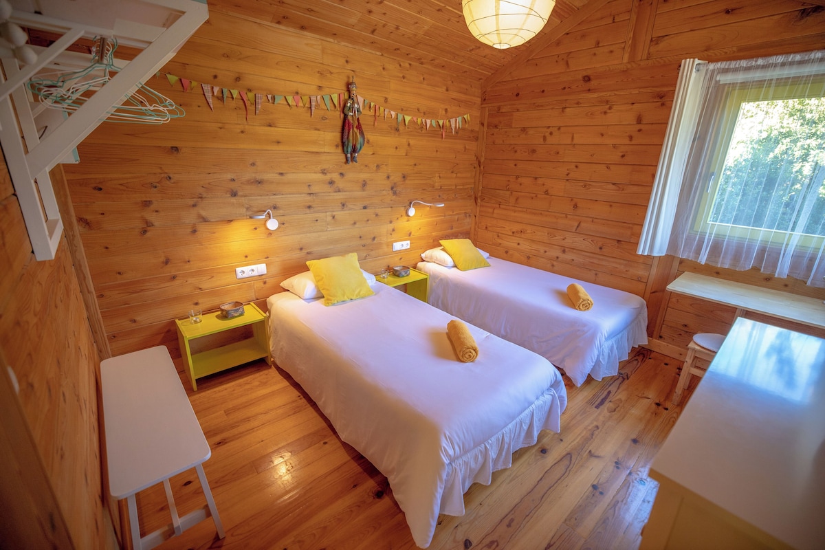 A wooden cabin room with two beds, a small table, and a window with curtains.