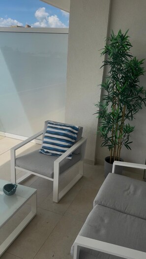 Ein Balkon mit einem weißen Stuhl, einem blau-weiß gestreiften Kissen, einem Blumentopf und einem grauen Sofa.