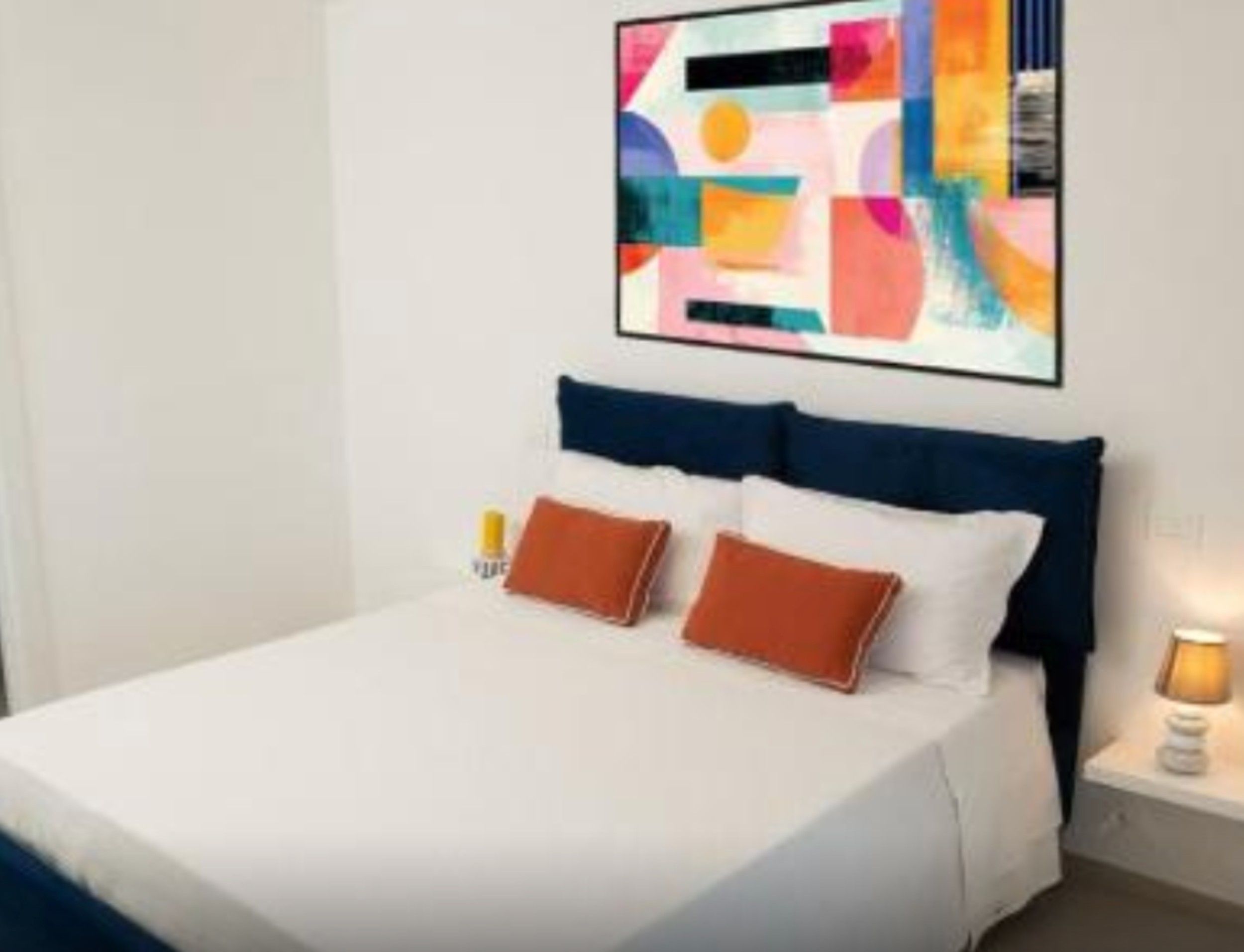 Ein Hotelzimmer mit Bett, Kissen, einem Nachttisch, einer Lampe und einem farbenfrohen, abstrakten Gemälde an der Wand.