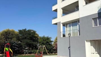 Un parque infantil con diversos juegos, incluyendo columpios y un tobogán, ubicado frente a un edificio moderno con balcones.