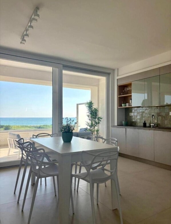 Eine moderne Küche mit einem großen Fenster, einem Essbereich mit weißen Stühlen und Blick aufs Meer.