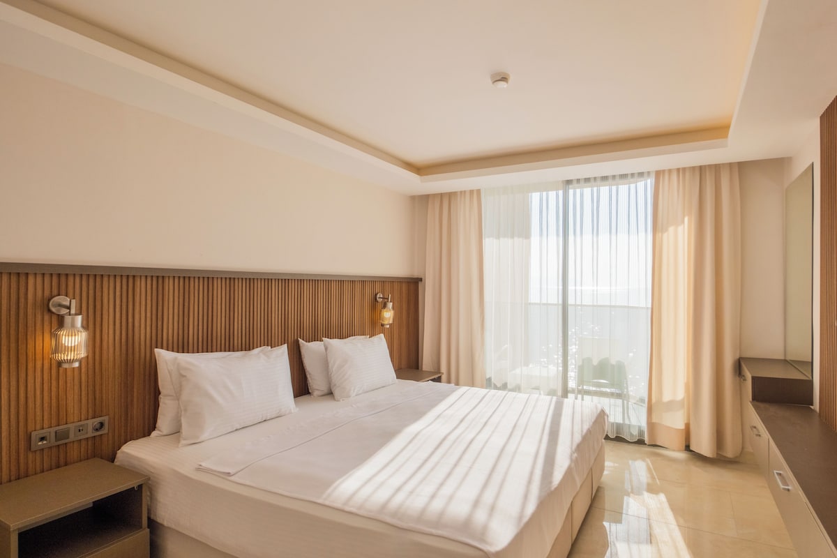 Une chambre d’hôtel avec un grand lit, une tête de lit en bois, une table de chevet, un bureau et une grande fenêtre avec des rideaux.