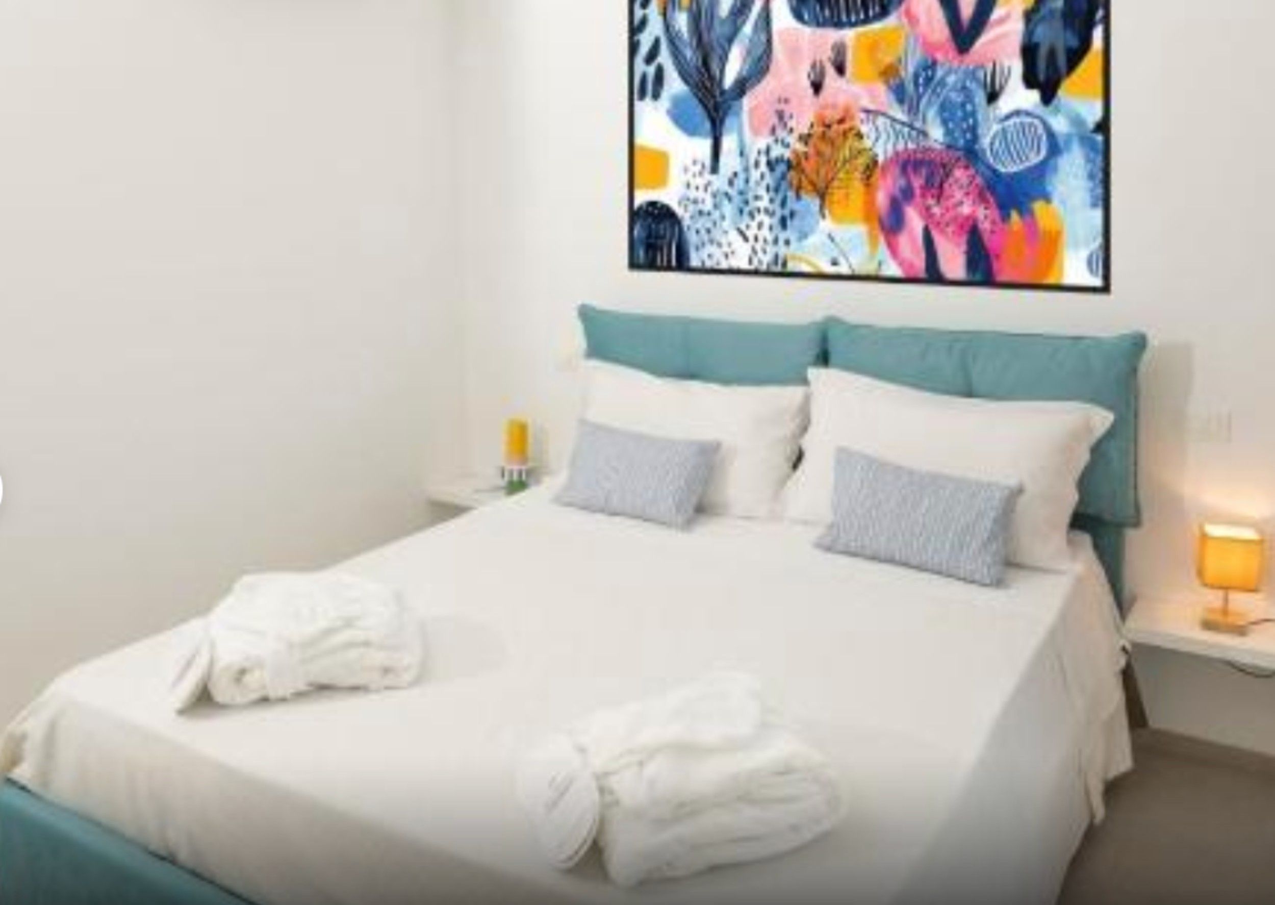 Una camera d'albergo con un letto, cuscini, un comodino, una lampada e un quadro astratto dai colori vivaci appeso alla parete.