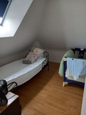 Enkeltseng med hvid madras og blomstret pude, et lille natbord med lampe, og et vindue med udsigt til himlen.