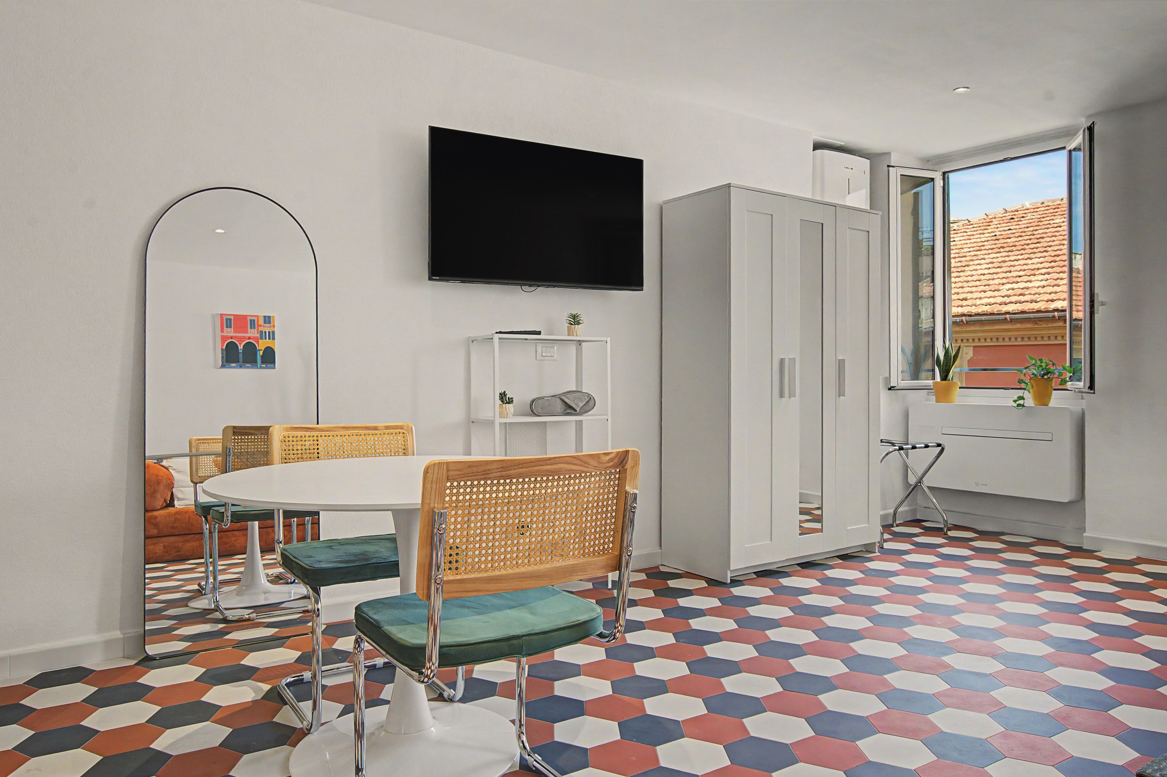 Ein Zimmer mit einem runden Esstisch, Stühlen, einem an der Wand befestigten Fernseher, einem Kleiderschrank und einem Fenster mit Blick auf Gebäude.