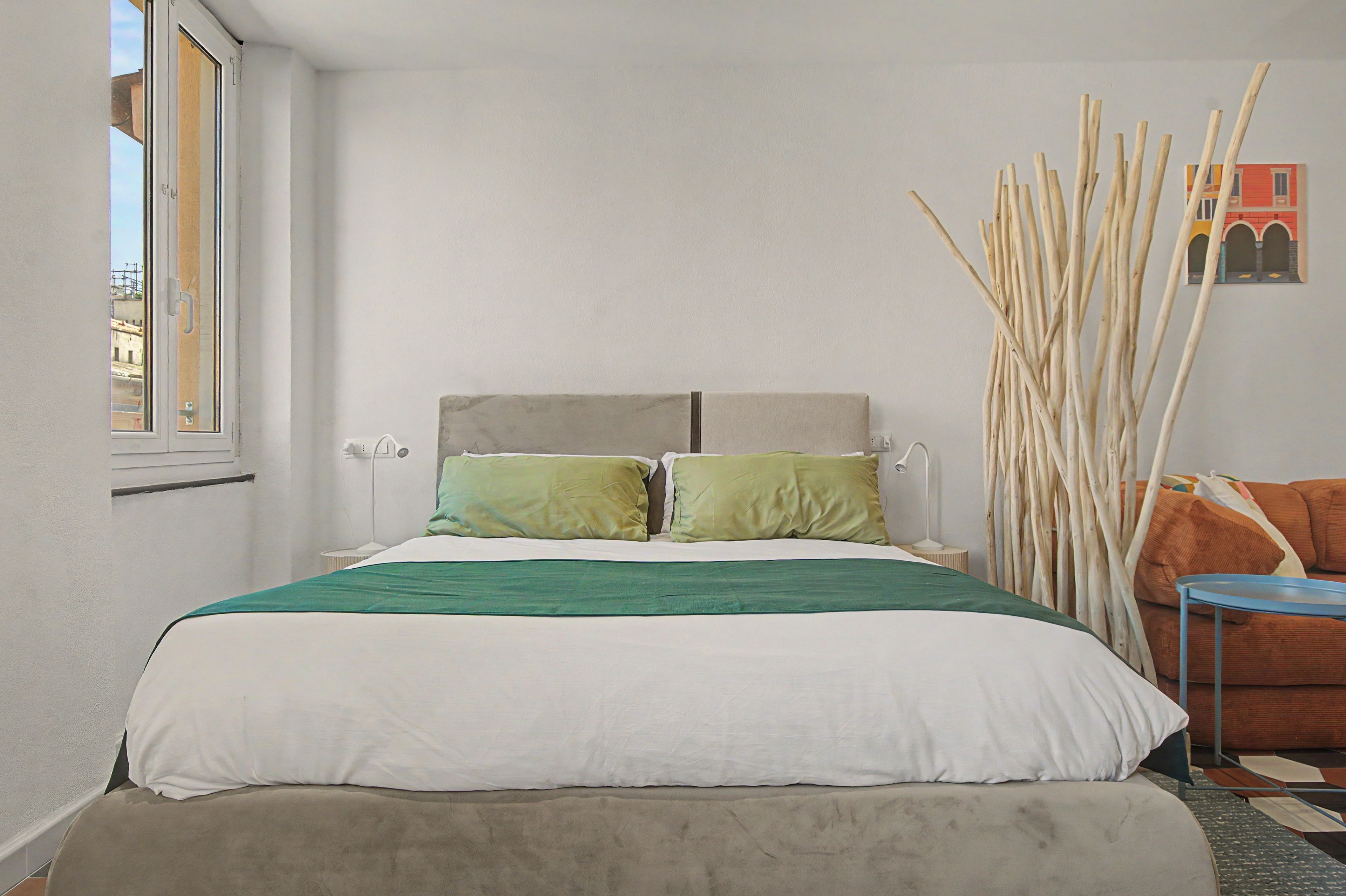 Une chambre à coucher avec un lit, des oreillers, une table de chevet, une lampe, une fenêtre et un tableau accroché au mur.