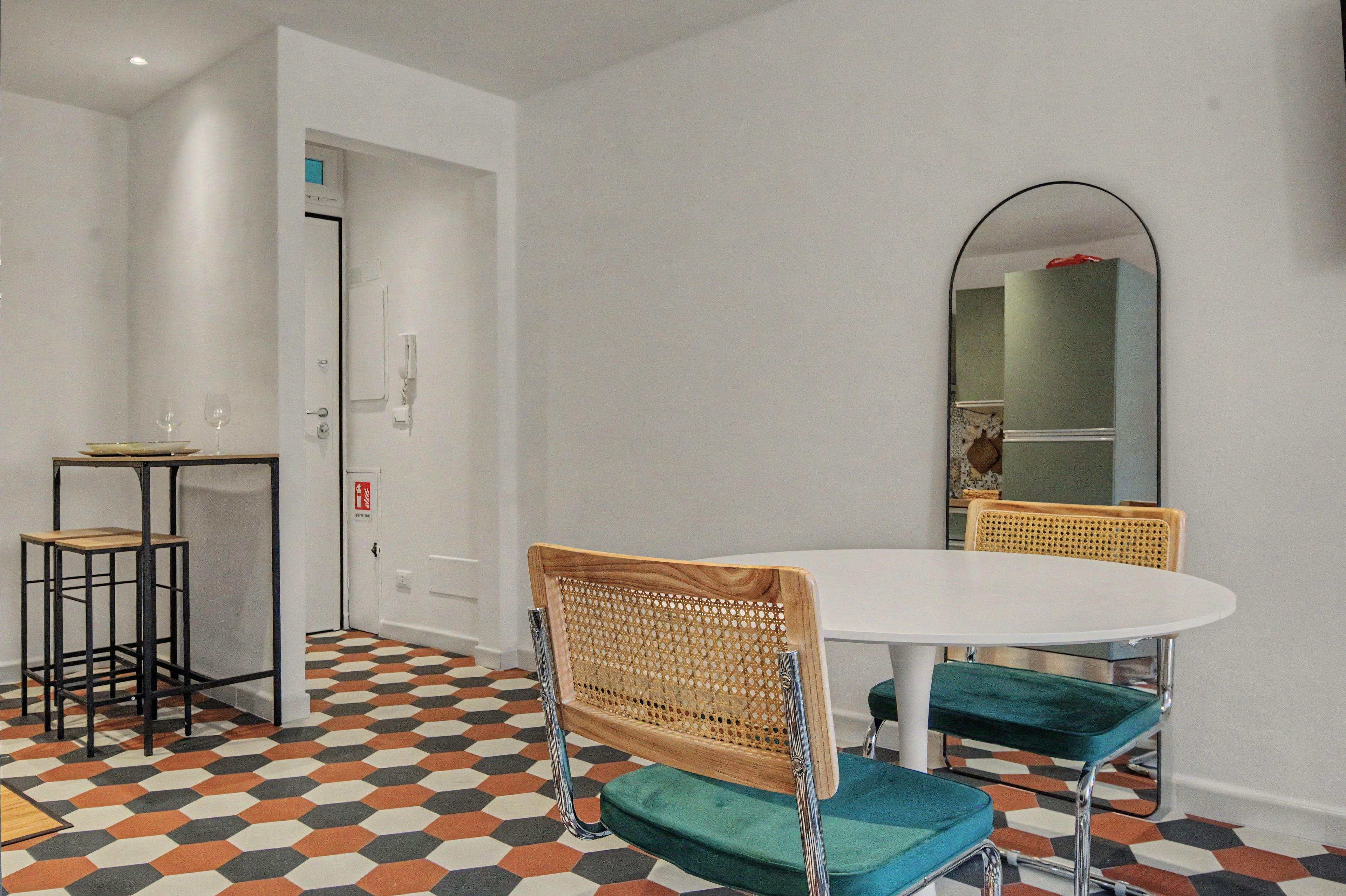 A dining area with a round table, two chairs, a small side table, and a mirror on the wall.