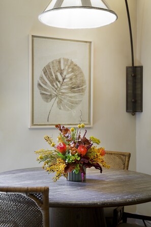 Uma mesa de jantar com um arranjo floral, uma obra de arte com folhas emolduradas na parede e um abajur.