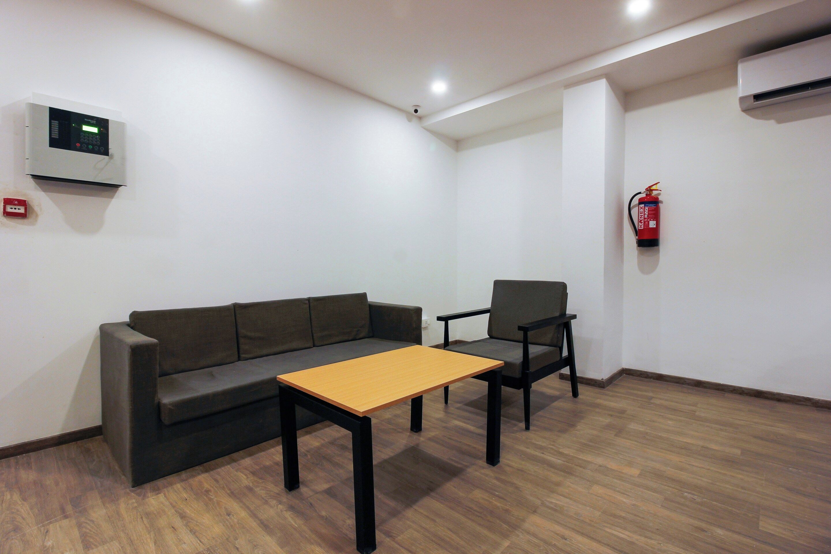 Quarto com sofá, poltrona, mesa pequena, extintor de incêndio e termostato na parede.