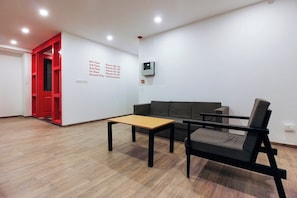 Una habitación amplia con piso de madera, una puerta roja, un sofá gris, una mesita y un panel de control fijado a la pared.