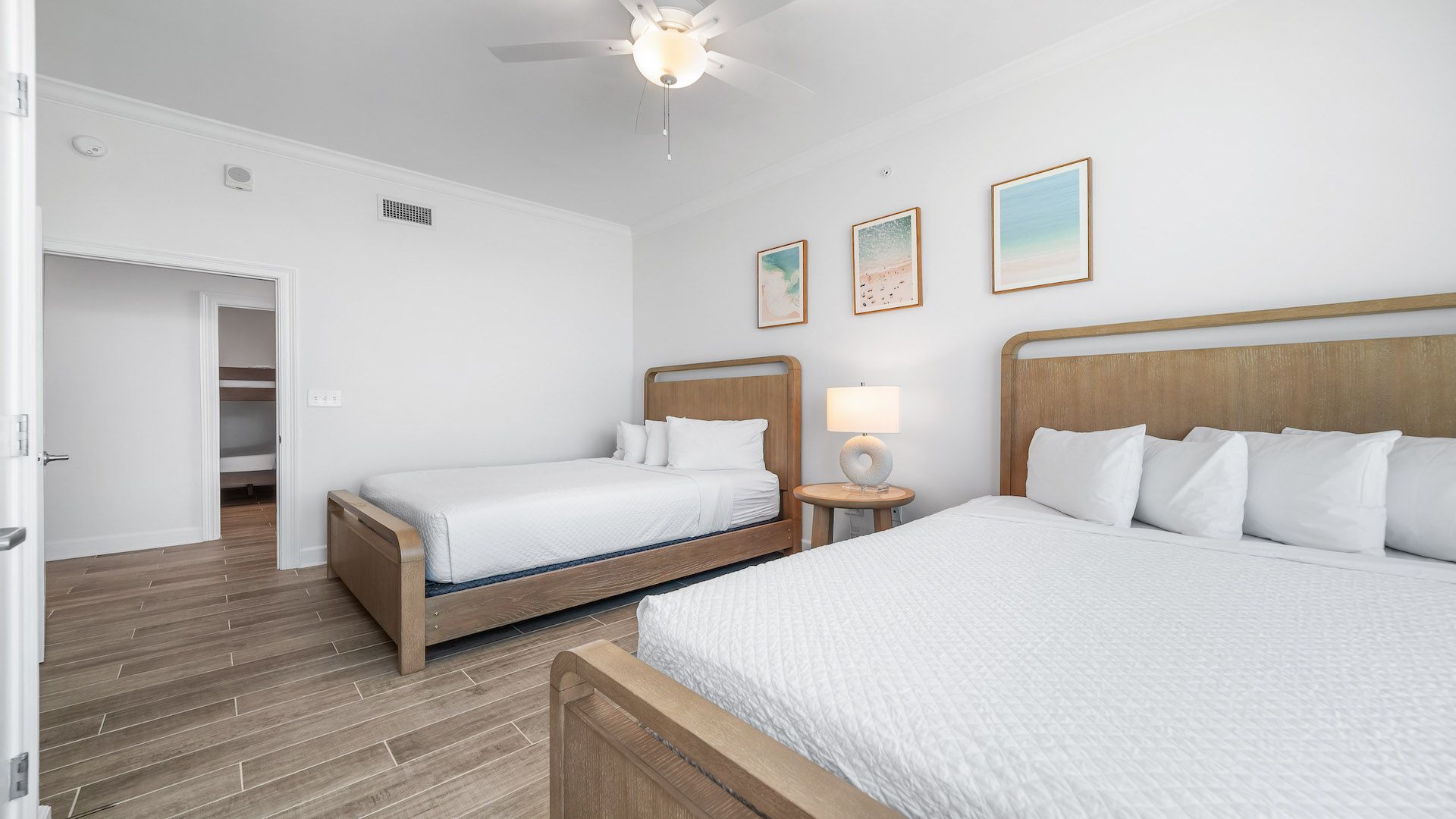 A hotel room with two beds, a wooden floor, a ceiling fan, and a door leading to a bathroom.