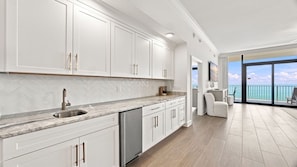 Une cuisine moderne avec des meubles blancs, un dosseret en carreaux disposés en chevron et une grande fenêtre donnant sur un balcon avec vue sur l’océan.