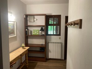 A small room with a wooden shelf unit, a radiator, a window with a view of greenery, and a small table with a vase.