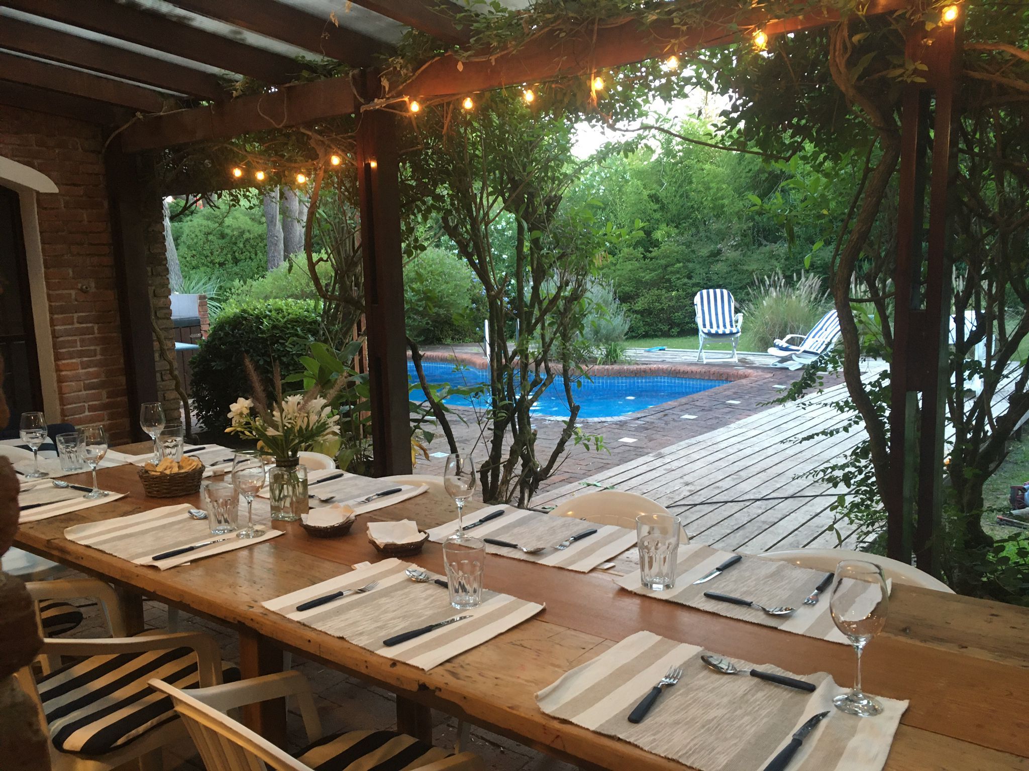 An outdoor dining area with a wooden table set for a meal, surrounded by greenery and a view of a swimming pool.