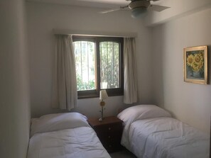 A room with two beds, a window with curtains, a ceiling fan, and a framed picture on the wall.