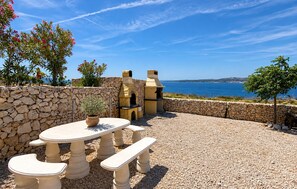 Un patio al aire libre con pared de piedra, mesa redonda, bancos y una parrilla, con vista a un cuerpo de agua.