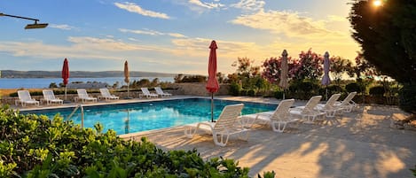 Zona de piscina con sillas de descanso y sombrillas, rodeada de vegetación y vistas al mar.