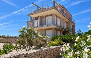 Un edificio residencial de varios pisos con balcones, un muro de piedra de contención y varios arbustos y flores en primer plano.
