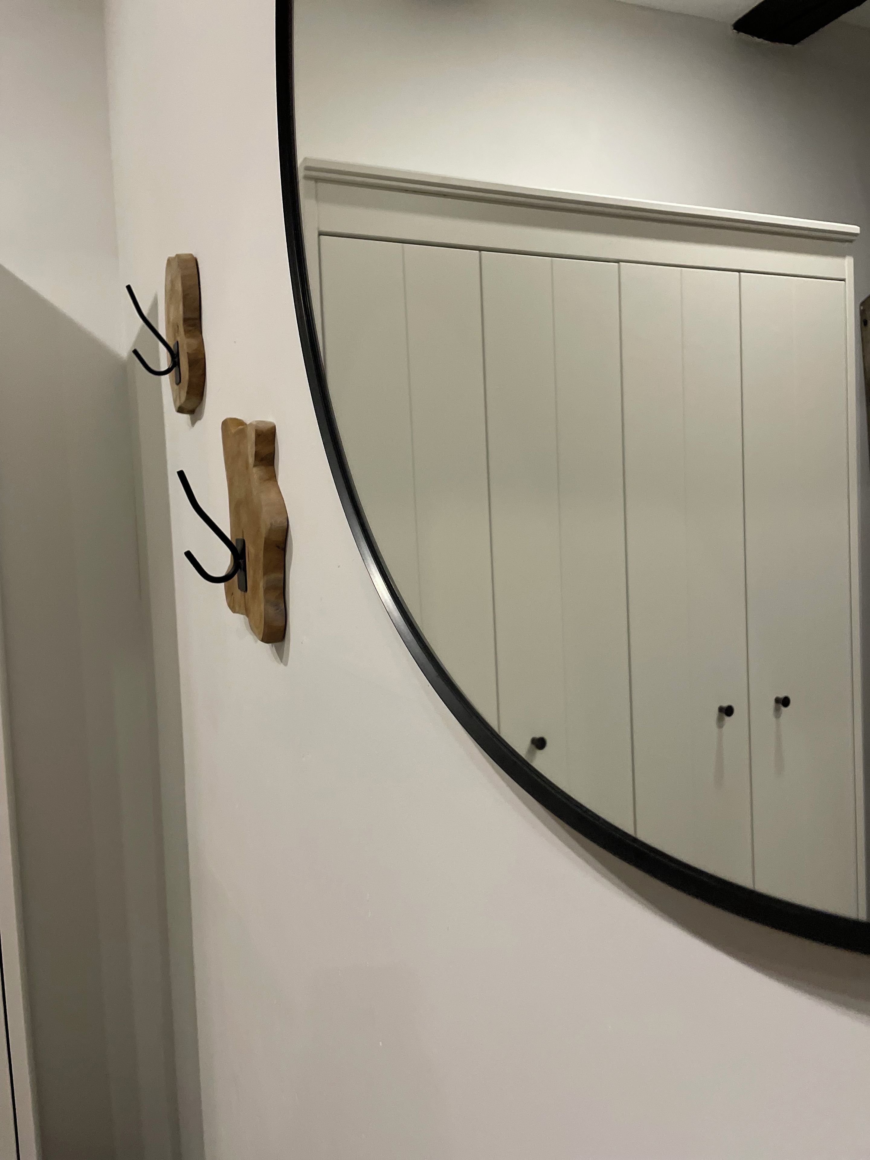 A room with a white wall, a round mirror, and two wooden wall hooks.