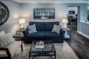 Ein modernes Wohnzimmer mit einem dunkelblauen Sofa, zwei Beistelltischen mit Lampen, einem Glastisch und einem Gemälde an der Wand. Im Hintergrund ist eine Küche zu sehen.