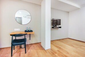 A room with a wooden desk, a chair, a round mirror, a television mounted on the wall, and a small shelf with bottles.