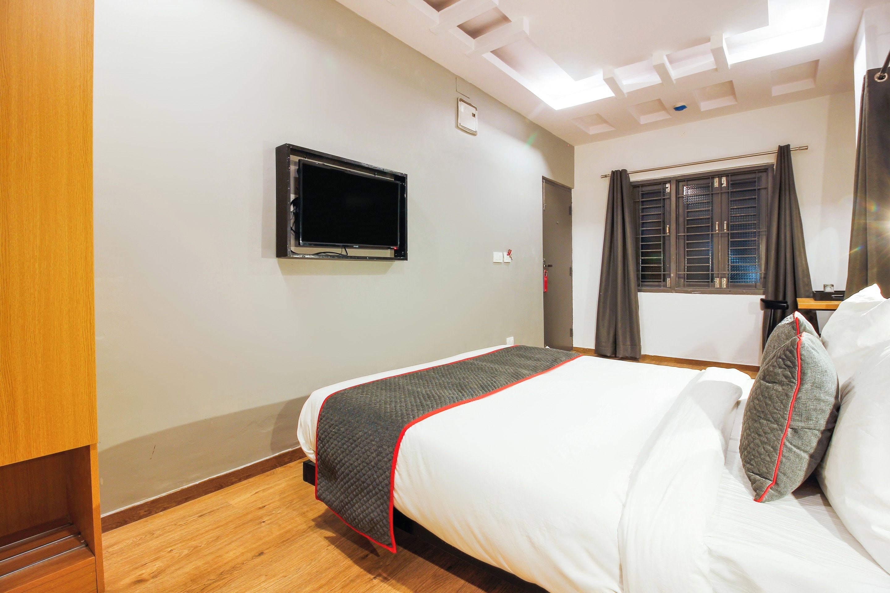 Une chambre d’hôtel avec un lit, un téléviseur fixé au mur, une fenêtre avec des rideaux et un plancher en bois.