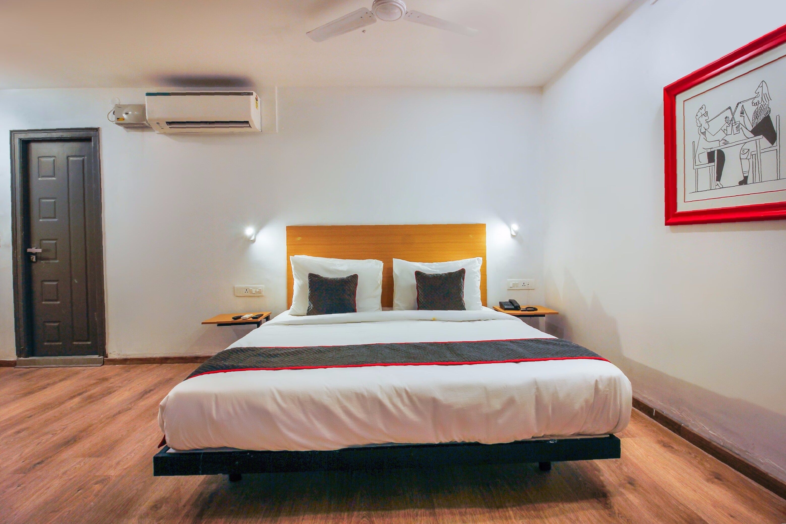 Een hotelkamer met een bed, nachtkastjes, een airconditioningapparaat, een deur en een lijst met een afbeelding aan de muur.